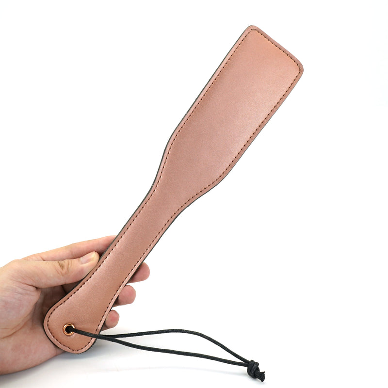Hand holding rose gold leather spanking paddle with wrist loop, ideal for BDSM play