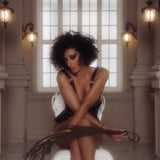 Elegant woman with curly hair in leather harness holding a leather Cat O' Nine Tails flogger from The Equestrian collection, seated in a room with classical architecture