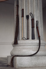 Assorted leather bondage whips and riding crops displayed against a classic stone column, featuring braided and smooth handles ideal for BDSM play