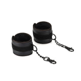 Black vegan leather wrist cuffs with metal chains and clips for bondage play, designed for comfort and safety in BDSM activities