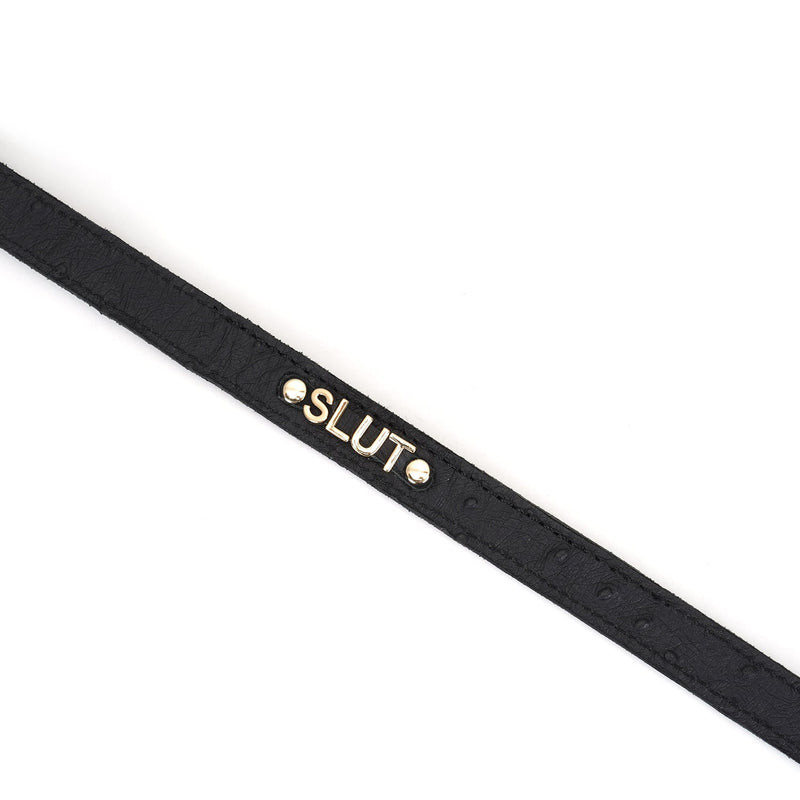 Black leather 'SLUT' choker from Demon's Kiss collection with ostrich skin pattern and gold lettering, close-up view