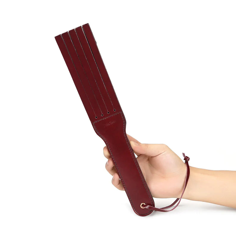 Hand holding wine red leather spanking paddle with slit details and hanging loop from the LIEBE SEELE bondage collection